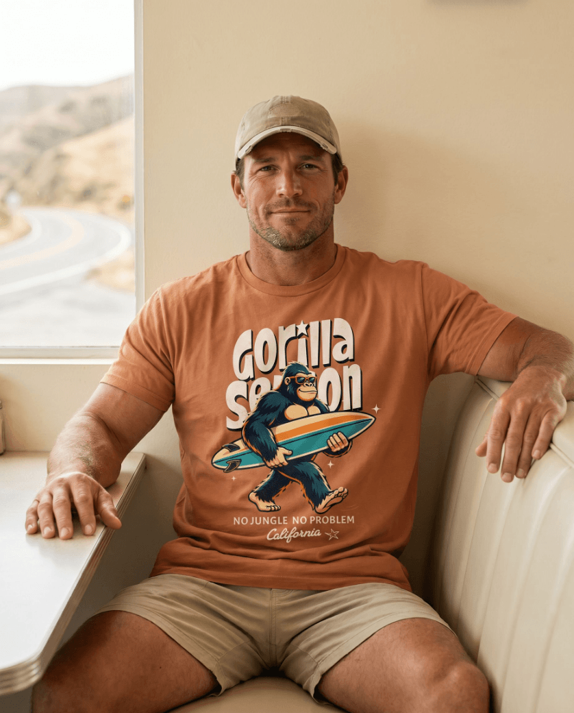 A man wearing a Gorilla Season Surf Tee – Yam, featuring a laid-back gorilla graphic, sitting indoors.