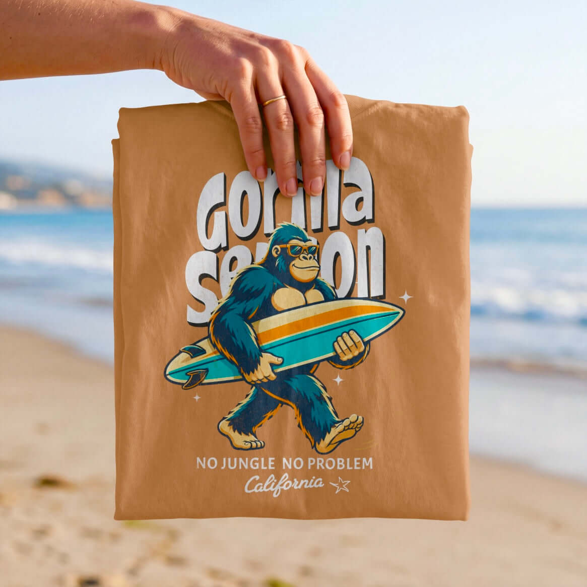Gorilla Season Surf Tee – Yam featuring a relaxed gorilla with a surfboard, beach backdrop, light brown color.