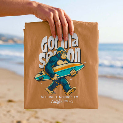 Gorilla Season Surf Tee – Yam featuring a relaxed gorilla with a surfboard, beach backdrop, light brown color.