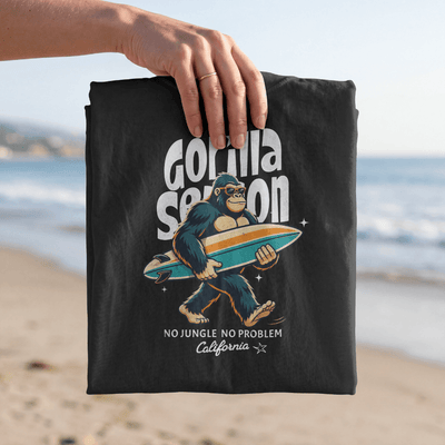 Gorilla Season Surf Tee – Black displayed on the beach, featuring a playful gorilla with a surfboard graphic.