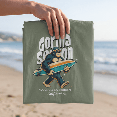Gorilla Season Surf Tee – Sage featuring a laid-back gorilla carrying a surfboard on the beach.