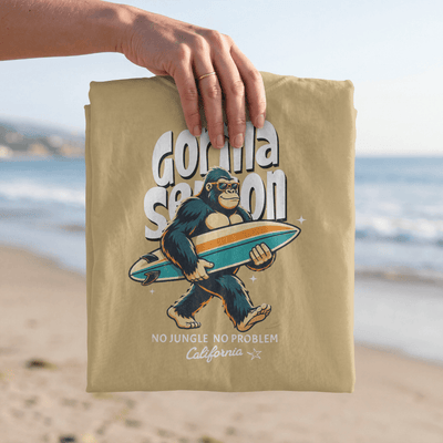 Gorilla Season Surf Tee – Mustard displayed on a beach with a bold gorilla graphic and surfboard.