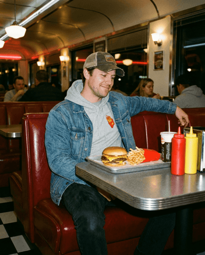 Man wearing a hoodie enjoys a burger and fries at a retro diner setting.