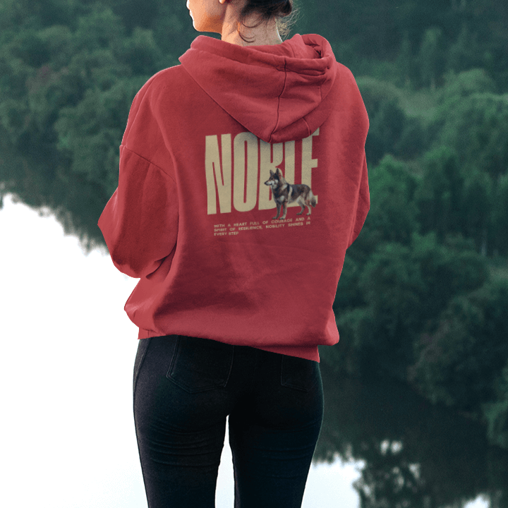 Woman in a red hoodie with "NOBLE" dog design, showcasing Nordic heritage by a scenic riverside.