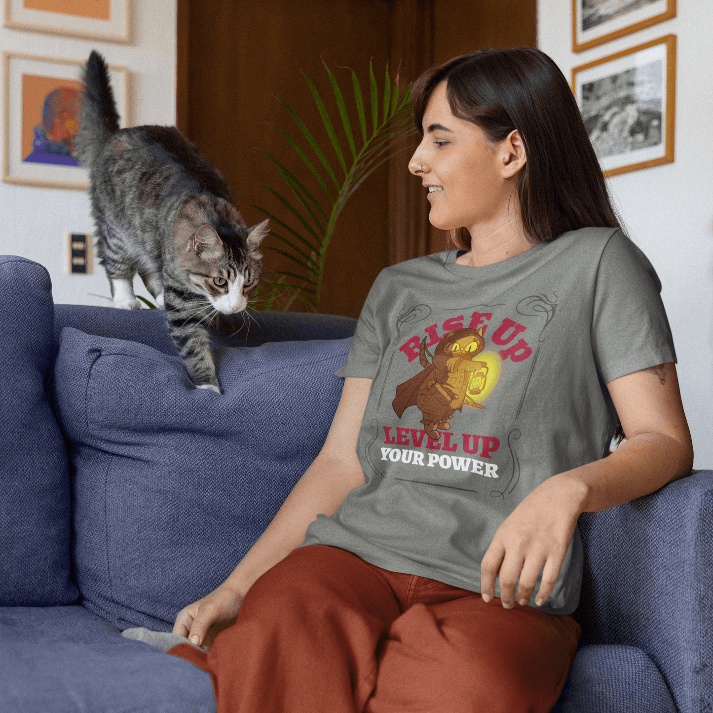 Mockup of a woman sitting on a couch wearing a gray T-shirt with a 'RISE UP, LEVEL UP YOUR POWER' warrior cat design, while interacting with a cat.