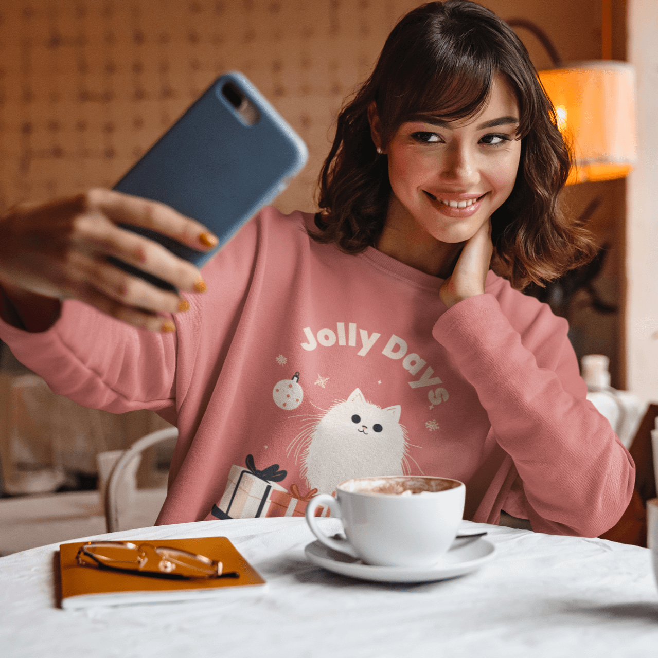 A young woman wearing a pink Christmas-themed sweatshirt sits in a cozy café, smiling while taking a selfie. The sweatshirt features the phrase 