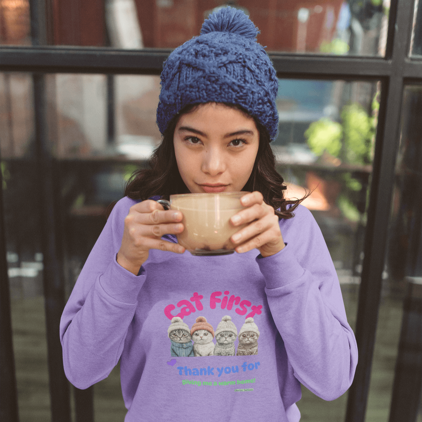 Young woman wearing a purple sweatshirt with a colorful design featuring four Scottish Fold cats in knitted hats and the text 'Cat First' and 'Thank you for giving me a warm home!' while holding a latte cup, sitting at a wooden table with a cafe window in