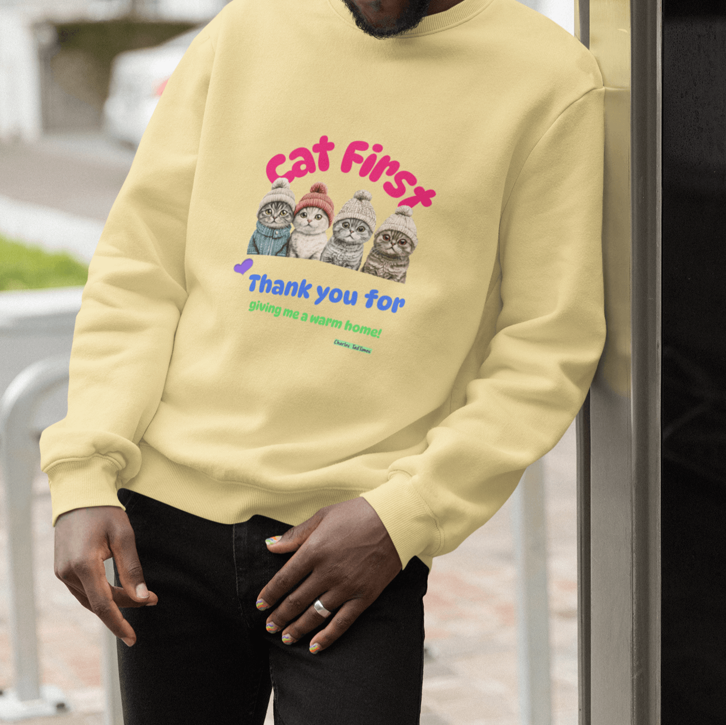 Smiling man wearing sunglasses and a yellow sweatshirt featuring a colorful design of four Scottish Fold cats in knitted hats, along with the text 'Cat First' and 'Thank you for giving me a warm home!' standing outdoors near a reflective surface.