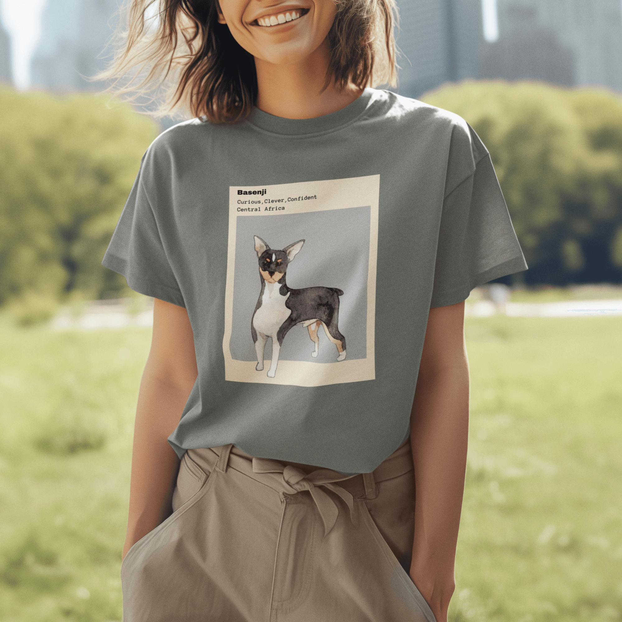 Smiling woman wearing a green T-shirt featuring a watercolor illustration of a Basenji dog with text describing its traits: 'Curious, Clever, Confident,' against a gray background, standing in a sunny park.