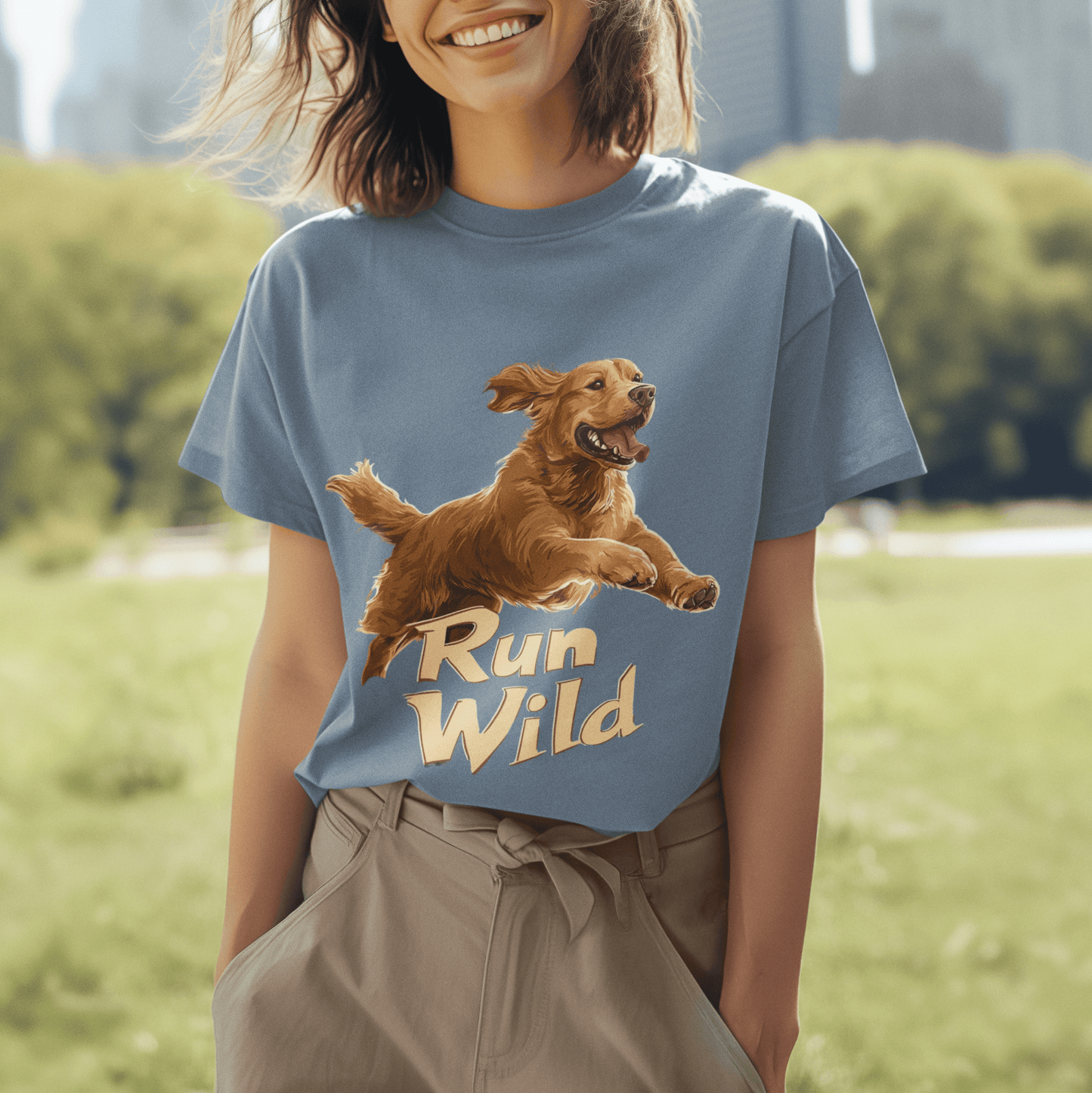 A smiling woman in a park wearing a blue T-shirt with a "Run Wild" design featuring a running Golden Retriever. The design highlights themes of freedom and energy, with a lush green field and a blurred city skyline in the background.