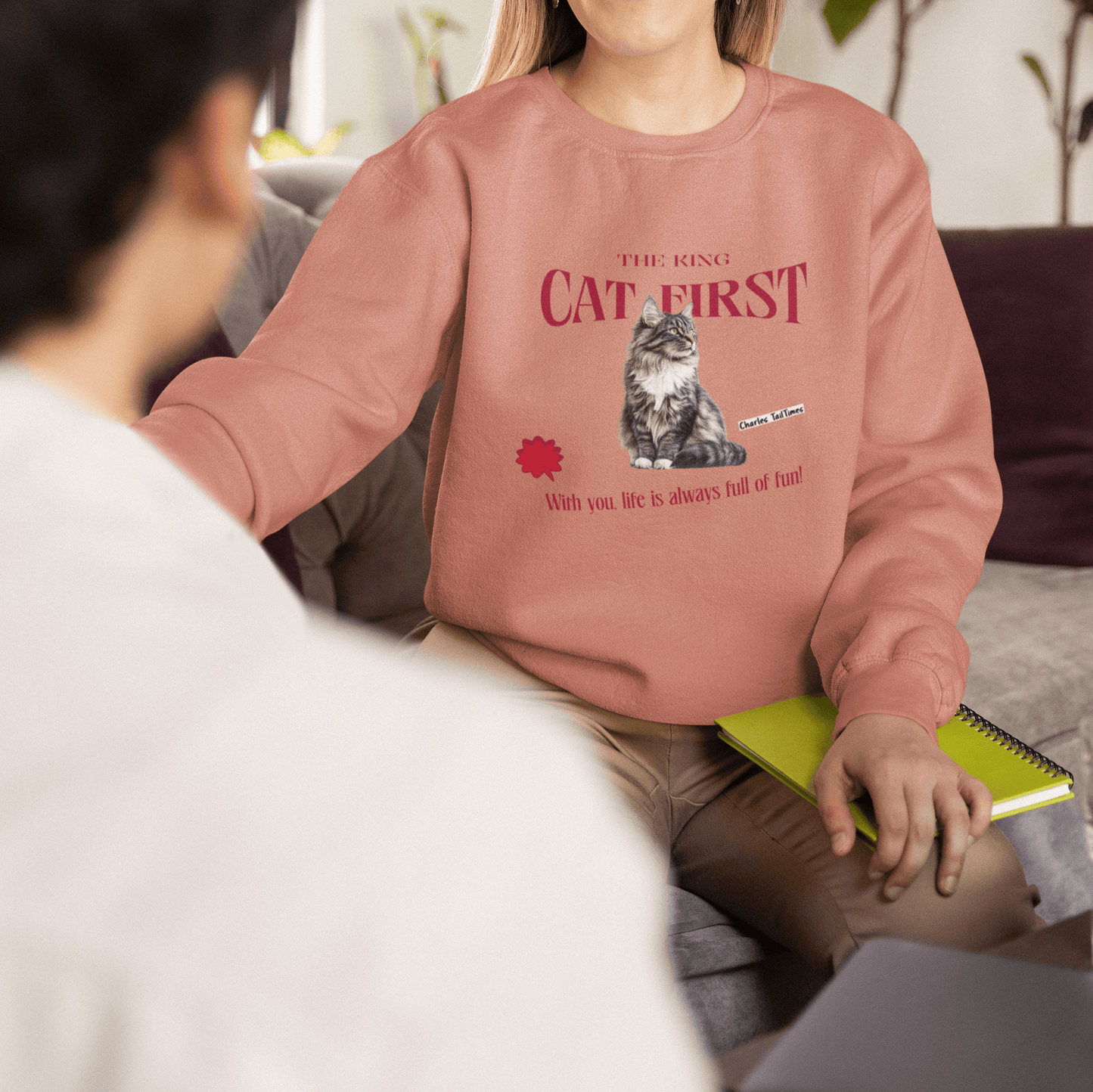 Peach sweatshirt featuring a Maine Coon cat graphic with the text "The King CAT FIRST" in red. A smiling woman holds a notebook while talking with a colleague.