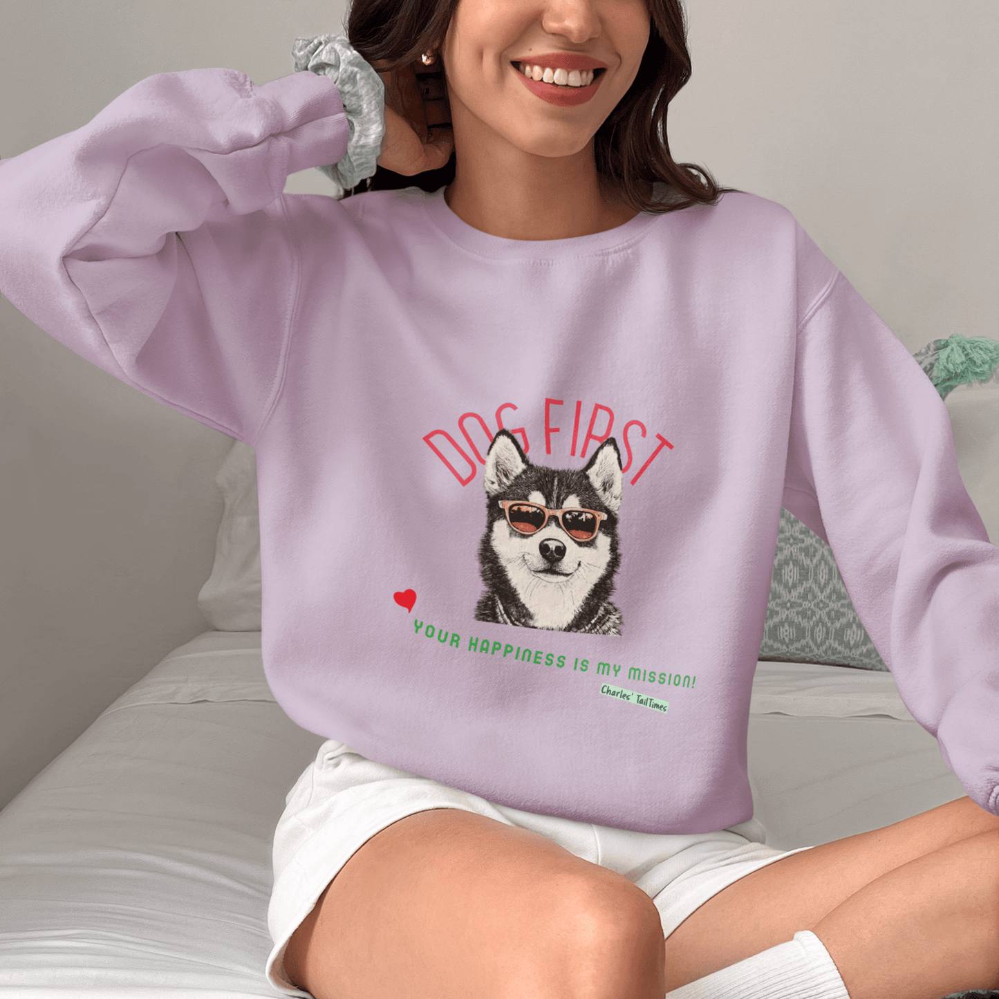 Female model wearing a purple "Dog First" illustration sweatshirt featuring a Husky design with sunglasses, set against a bed background.