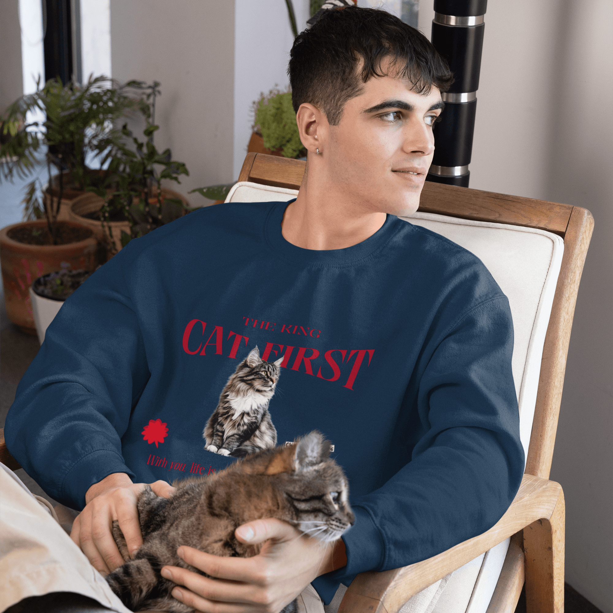 A man sitting on a chair, wearing a navy blue sweatshirt featuring a Maine Coon cat illustration with the text 