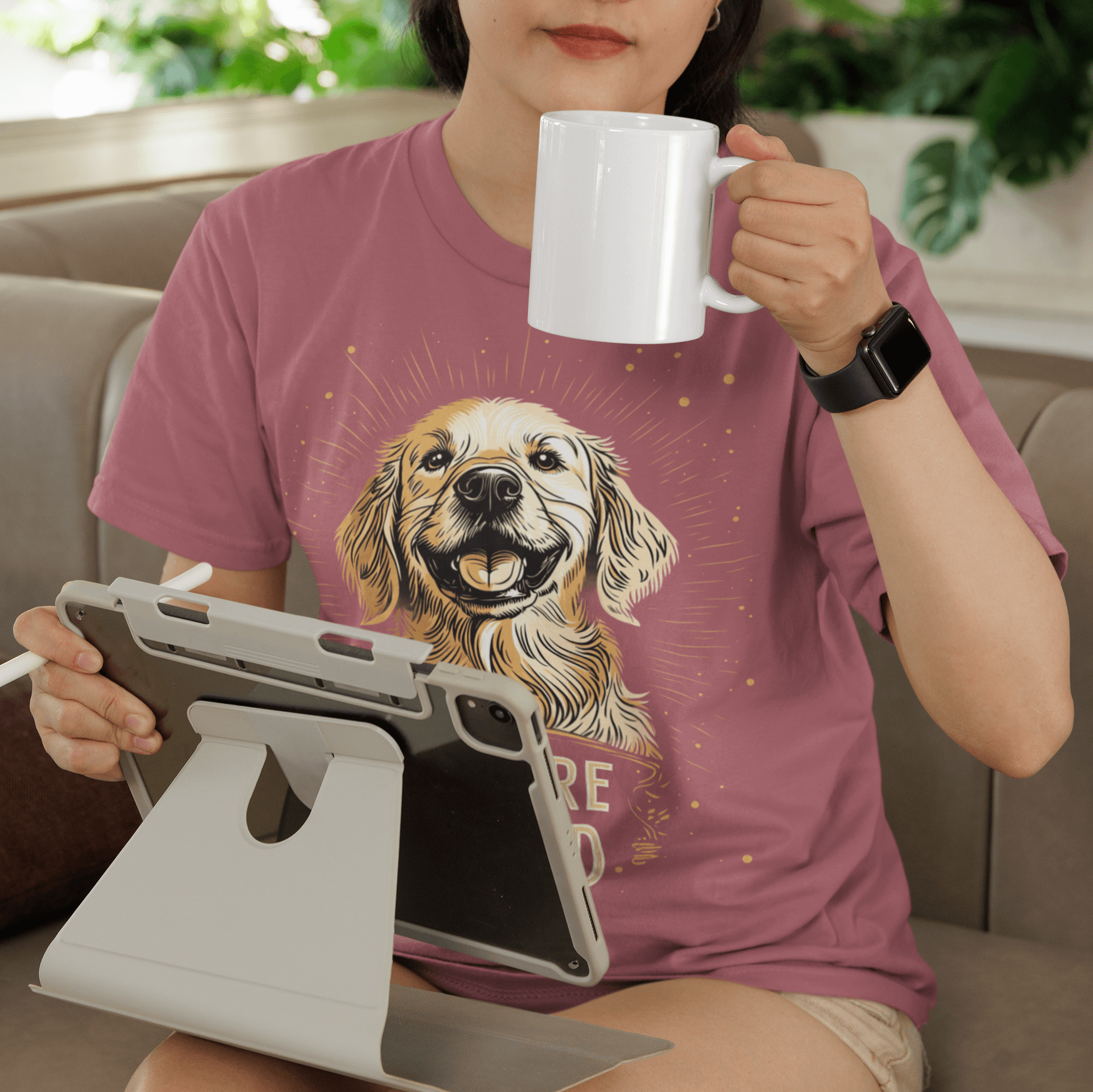 Woman sitting on a couch wearing a pink round-neck T-shirt featuring a smiling Golden Retriever illustration and the text 