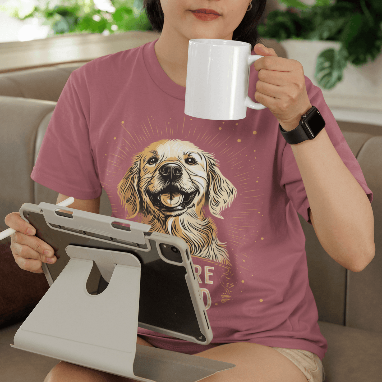 Woman sitting on a couch wearing a pink round-neck T-shirt featuring a smiling Golden Retriever illustration and the text "YOU'RE LOVED," holding a white coffee mug in one hand and using a tablet with the other, with lush greenery in the background for a 