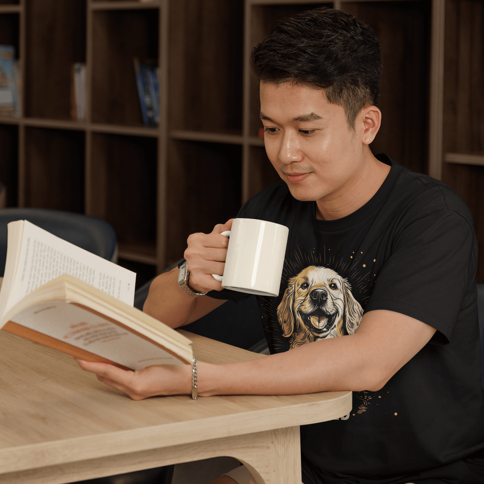 Man reading at a table, holding a white mug, wearing a black T-shirt with a Golden Retriever design and the text 