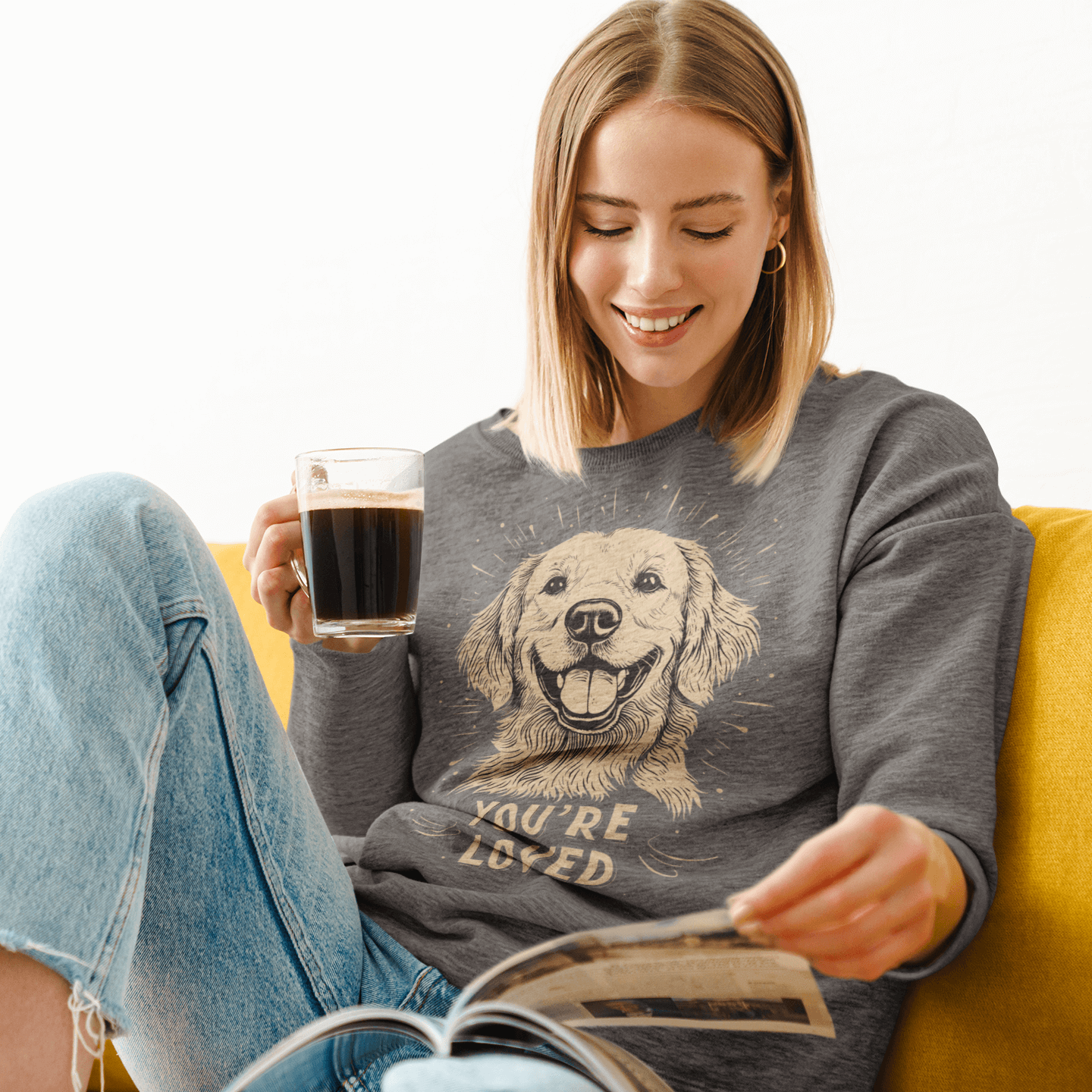 A woman sitting on a yellow couch wears a gray crewneck sweatshirt, holding a coffee cup while reading a magazine. The sweatshirt features an illustration of a smiling golden retriever and the text 