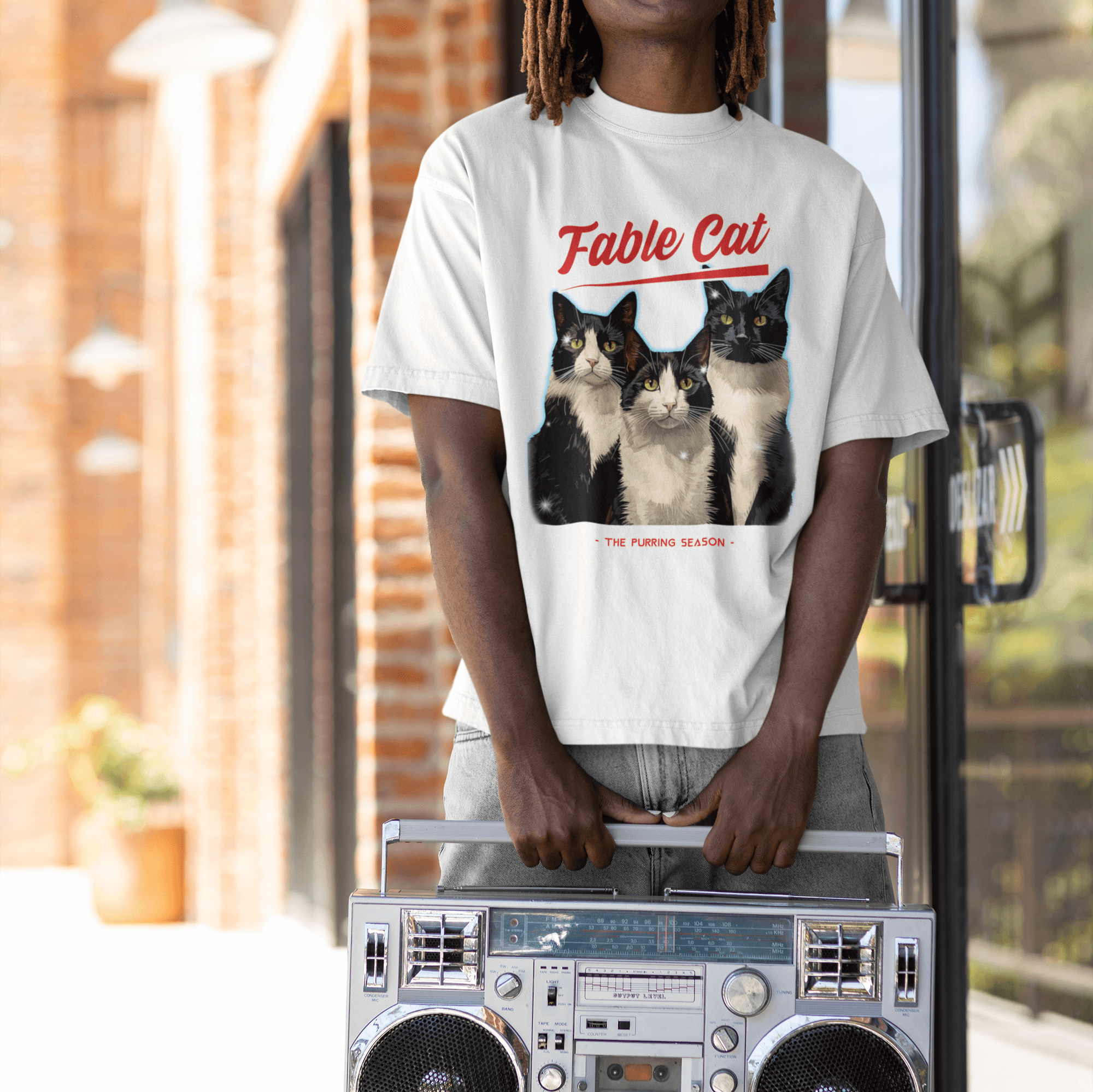 A man wearing a white T-shirt featuring an illustration of three tuxedo cats with the text 'Fable Cat' in red bold typography above them and the subtitle 'The Purring Season' below, standing outside holding a retro boombox.