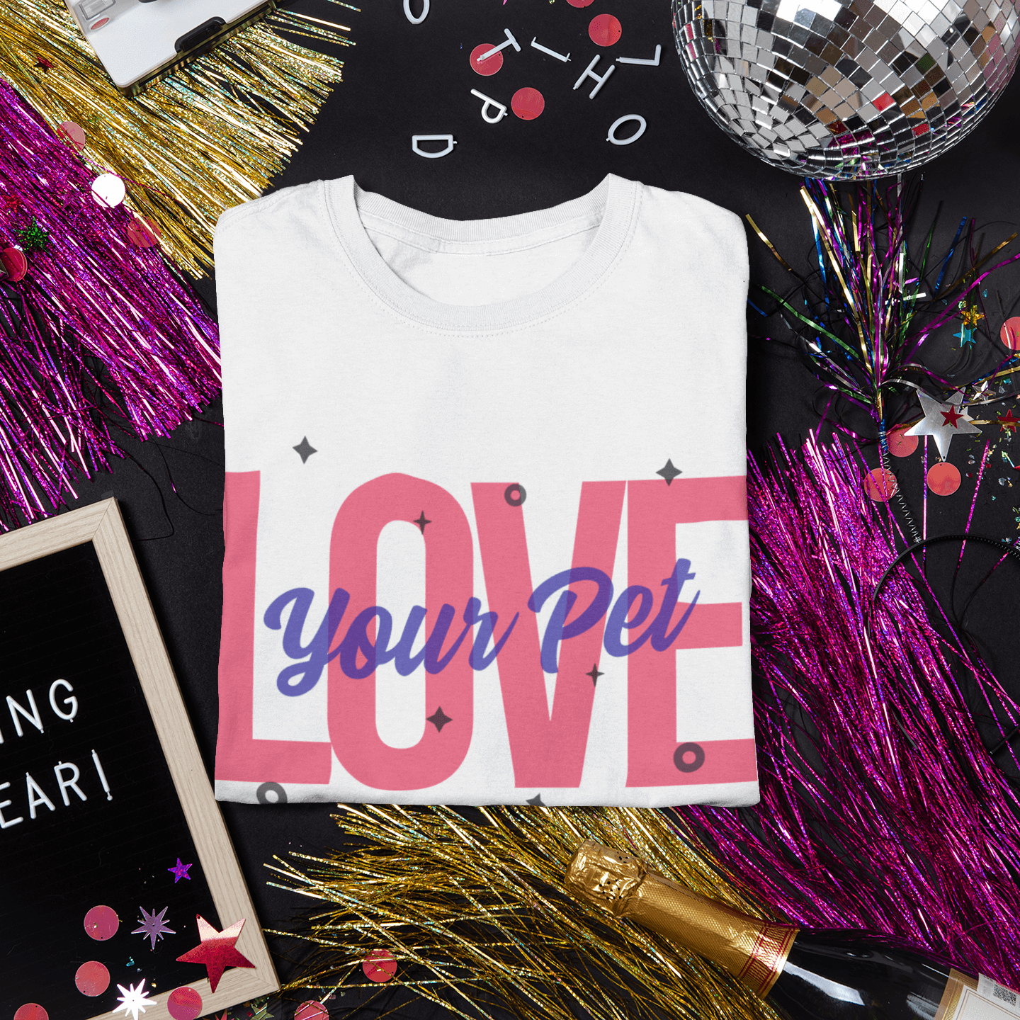 Folded white T-shirt with a bold 'Love Your Pet' design in pink and blue, displayed among sparkling New Year’s party decorations, including disco balls, confetti, and a champagne bottle.