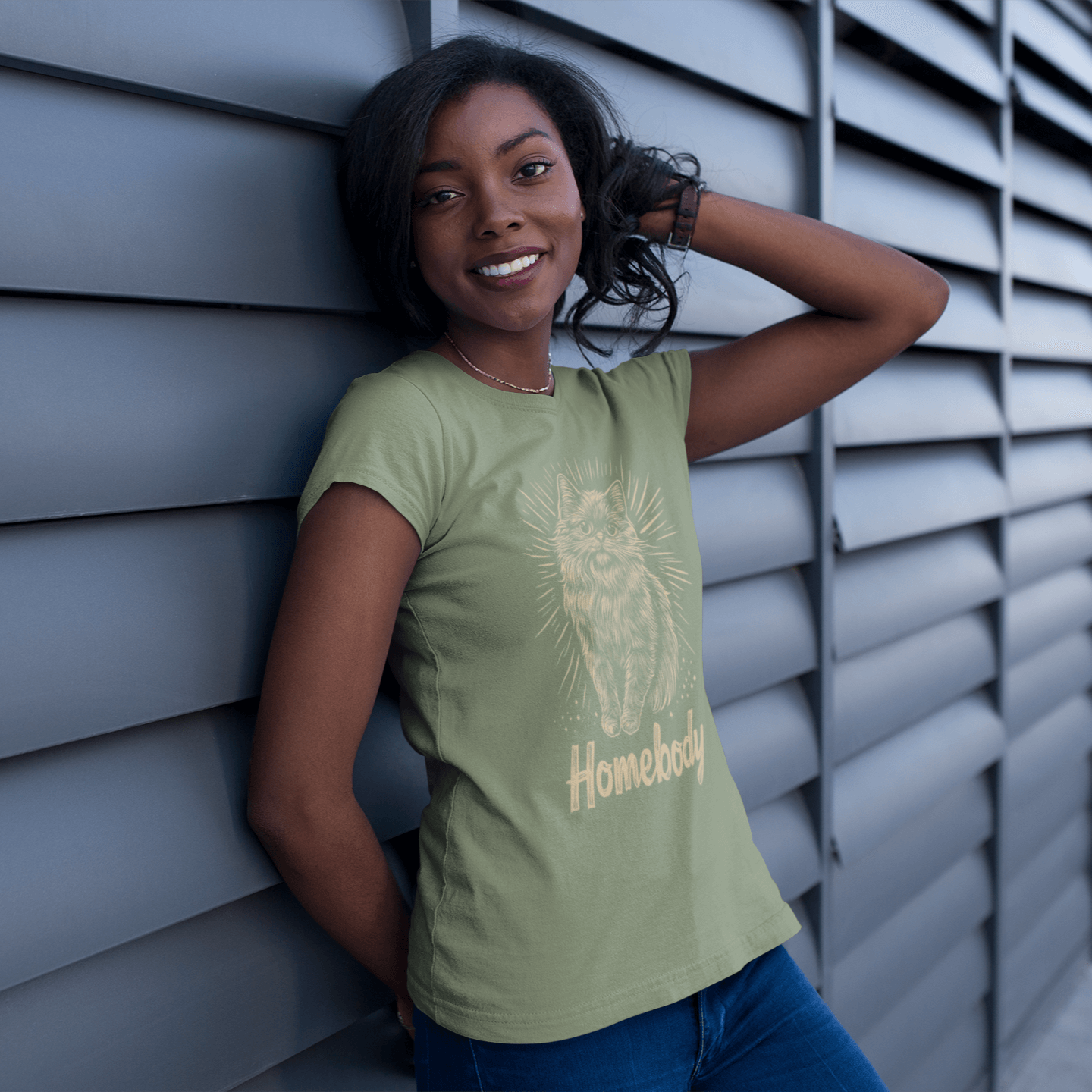 A model wearing a green T-shirt featuring a Persian cat illustration and the text 'Homebody,' leaning against a metal wall and smiling, showcasing a comfortable and confident style.