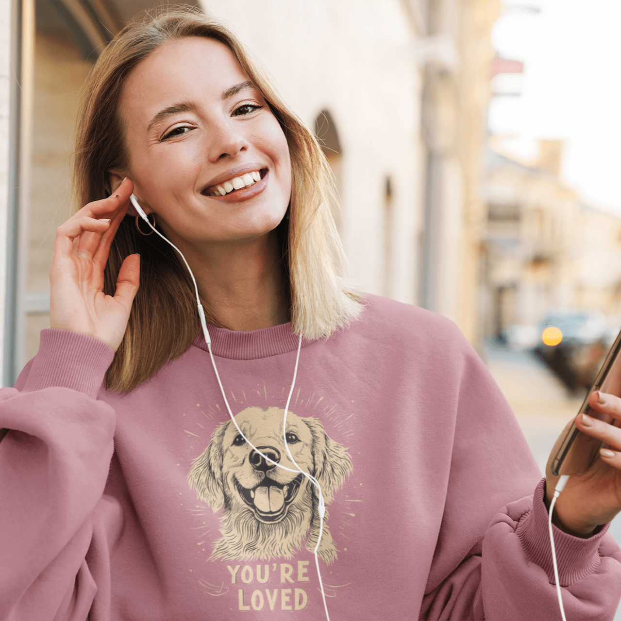 A smiling woman by the street wears a light pink crewneck sweatshirt, listening to earphones and holding a phone. The sweatshirt features a cheerful golden retriever illustration and the text 