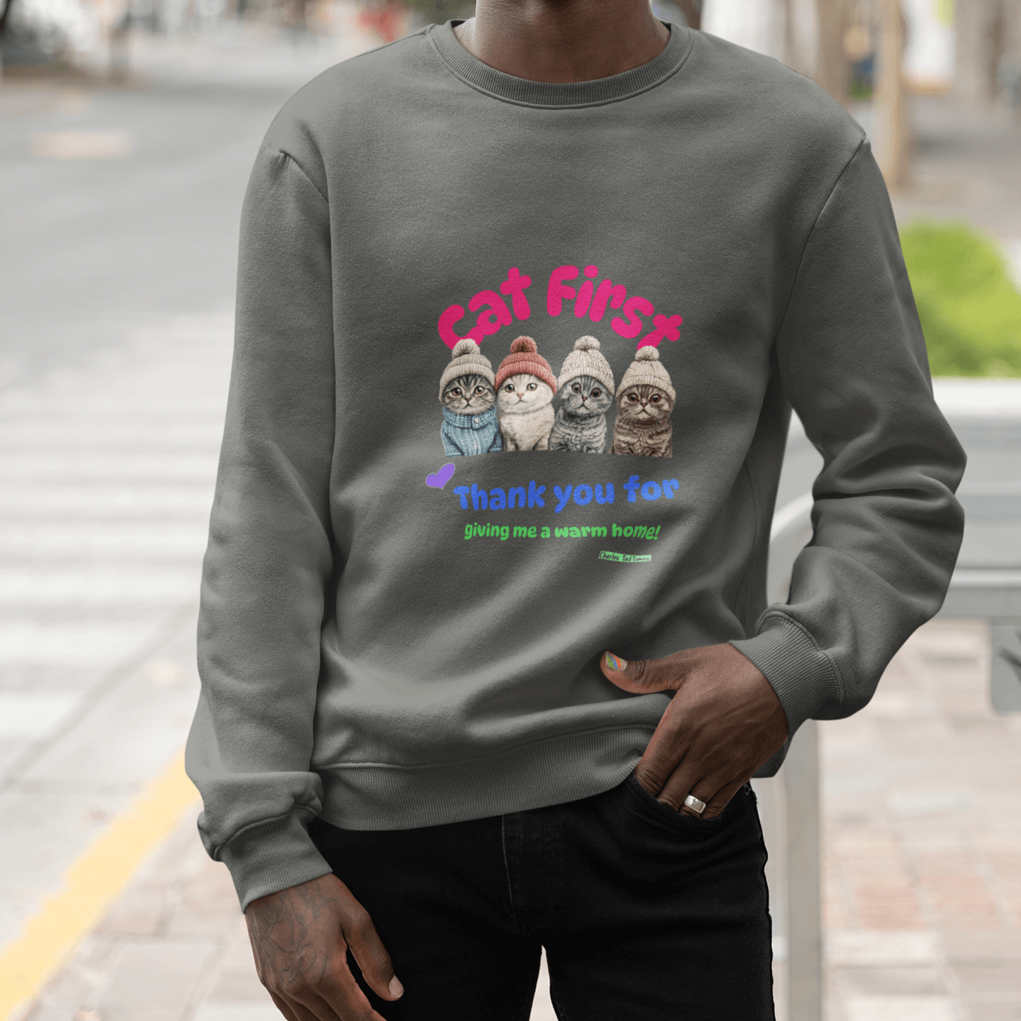 Happy man wearing sunglasses and a gray sweatshirt with a colorful cat-themed design featuring four Scottish Fold cats in knitted hats, alongside the text 'Cat First' and 'Thank you for giving me a warm home!' while standing outdoors.