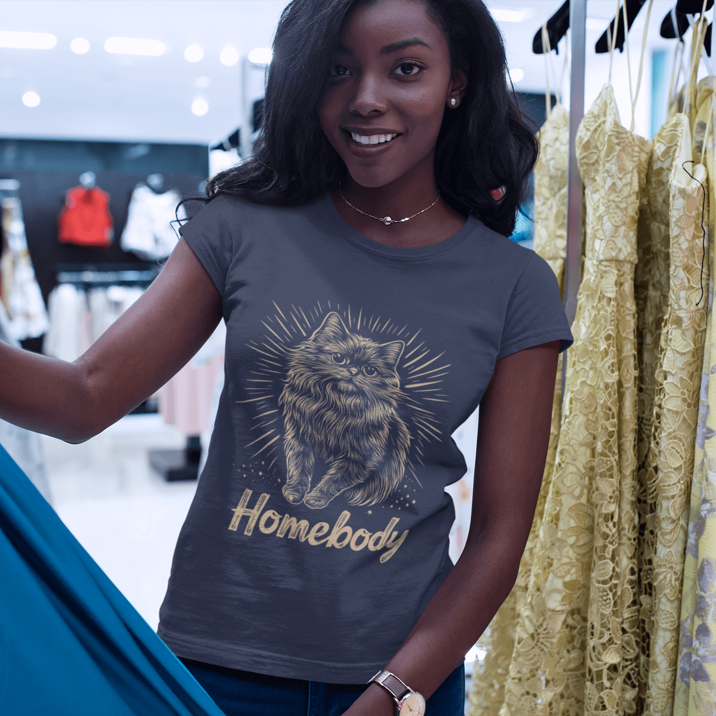 A model wearing a gray T-shirt featuring a Persian cat illustration and the text 'Homebody,' browsing clothes in a store, showcasing a relaxed and comfortable shopping moment.