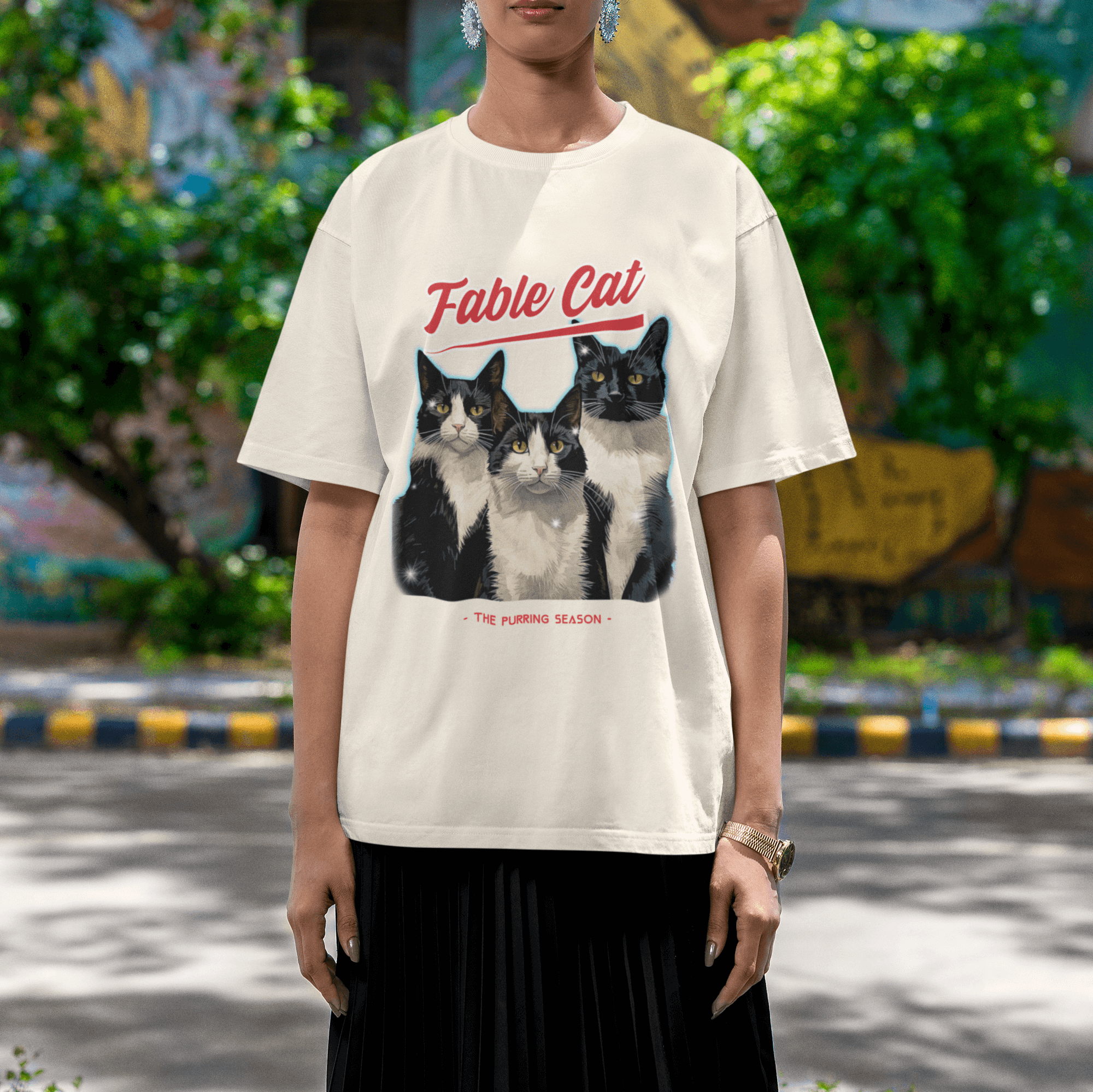 Woman standing outdoors wearing an oversized cream T-shirt with an illustration of three tuxedo cats, red 'Fable Cat' text, and the tagline 'The Purring Season.