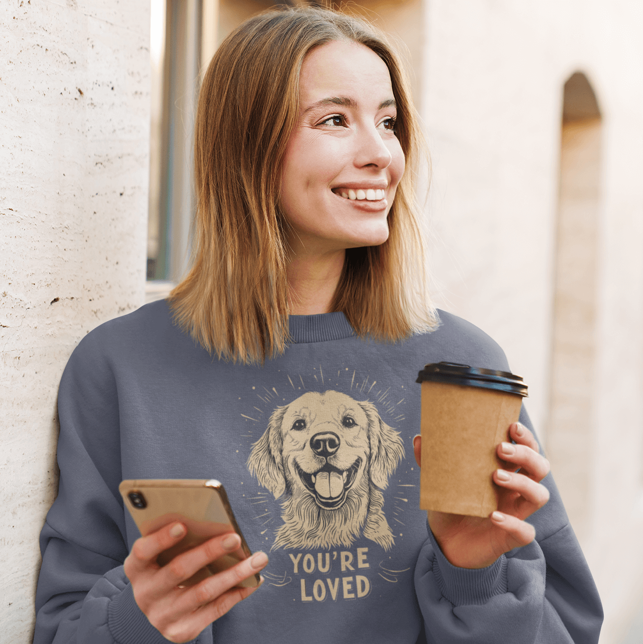 A woman standing by the street wears a light blue crewneck sweatshirt, holding a smartphone and a coffee cup while smiling. The sweatshirt features an illustration of a cheerful golden retriever and the text 