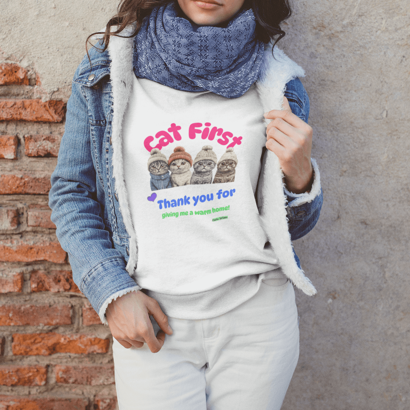 Young woman wearing a white sweatshirt with an illustration of four Scottish Fold cats in knitted hats and colorful text 'Cat First' and 'Thank you for giving me a warm home!' layered under a denim jacket, standing against a rustic brick wall.