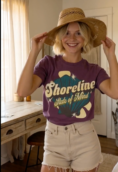 Woman wearing a Shoreline State of Mind Tee – Berry, smiling and holding a straw hat.