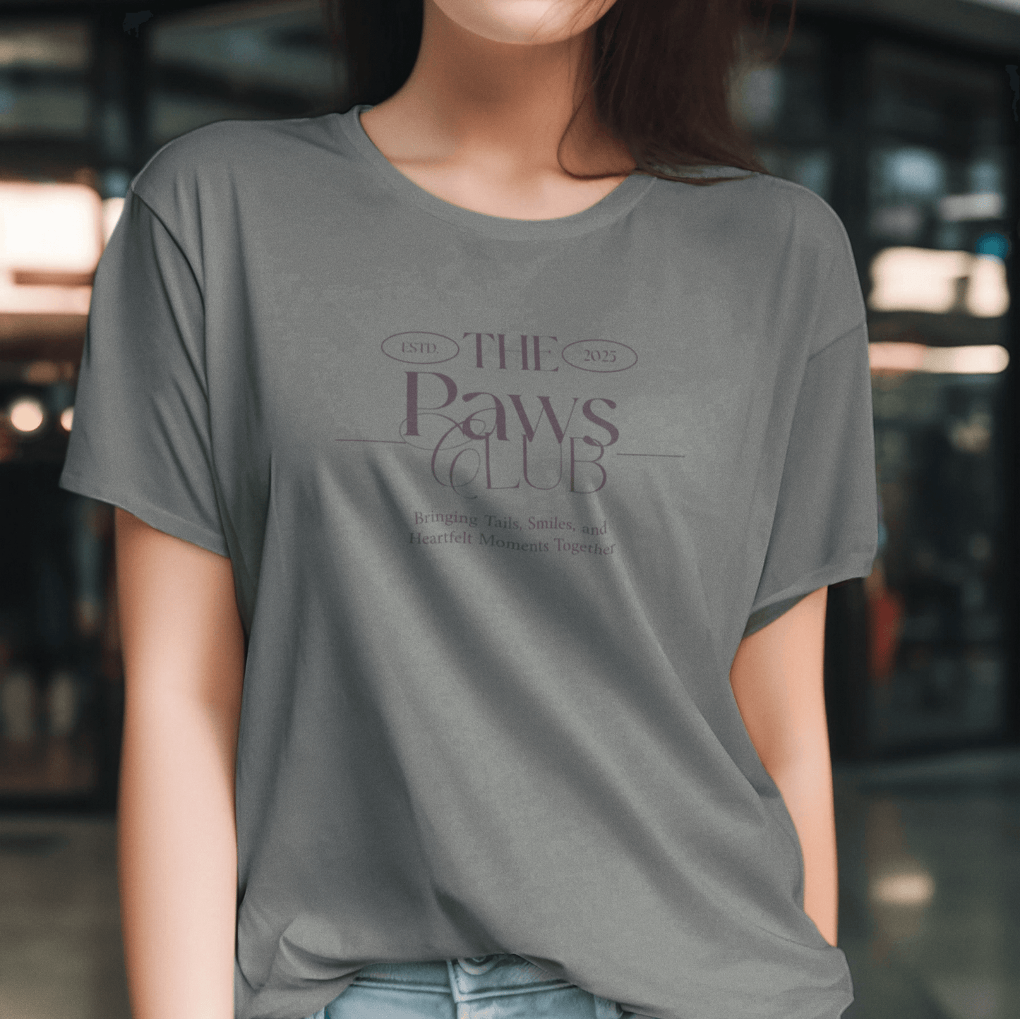 Stylish woman wearing a gray T-shirt featuring the text 'The Paws Club,' established in 2025, with the tagline 'Bringing Tails, Smiles, and Heartfelt Moments Together,' set against an urban background.