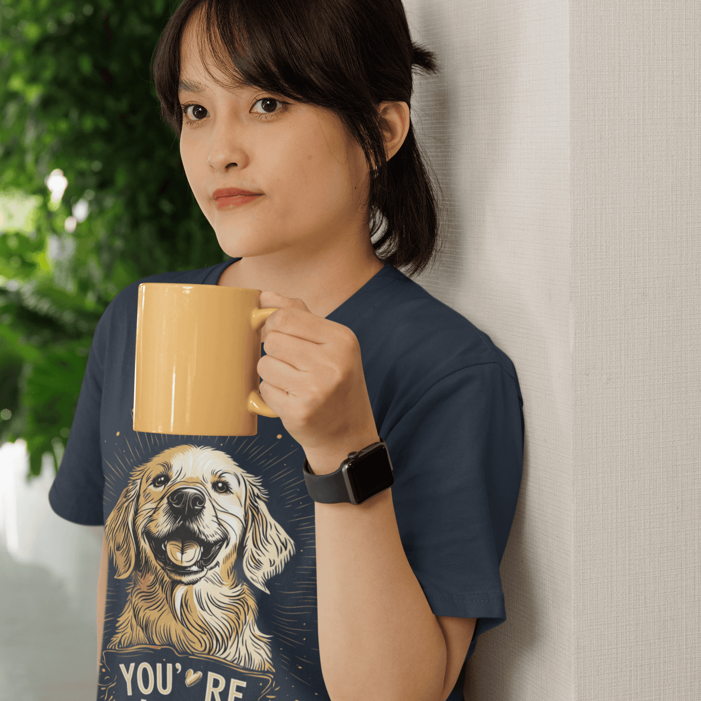 Alt text: Woman holding a yellow coffee mug, wearing a navy blue T-shirt with a smiling Golden Retriever illustration and the text "YOU'RE LOVED," exuding warmth and positivity indoors.