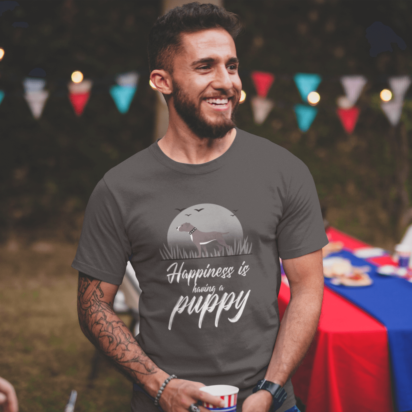 Smiling man wearing a dark gray T-shirt with a Pitbull design, standing at an outdoor BBQ with festive decorations and lights in the background. The shirt features the text 'Happiness is having a puppy' in white cursive font under a full moon illustration