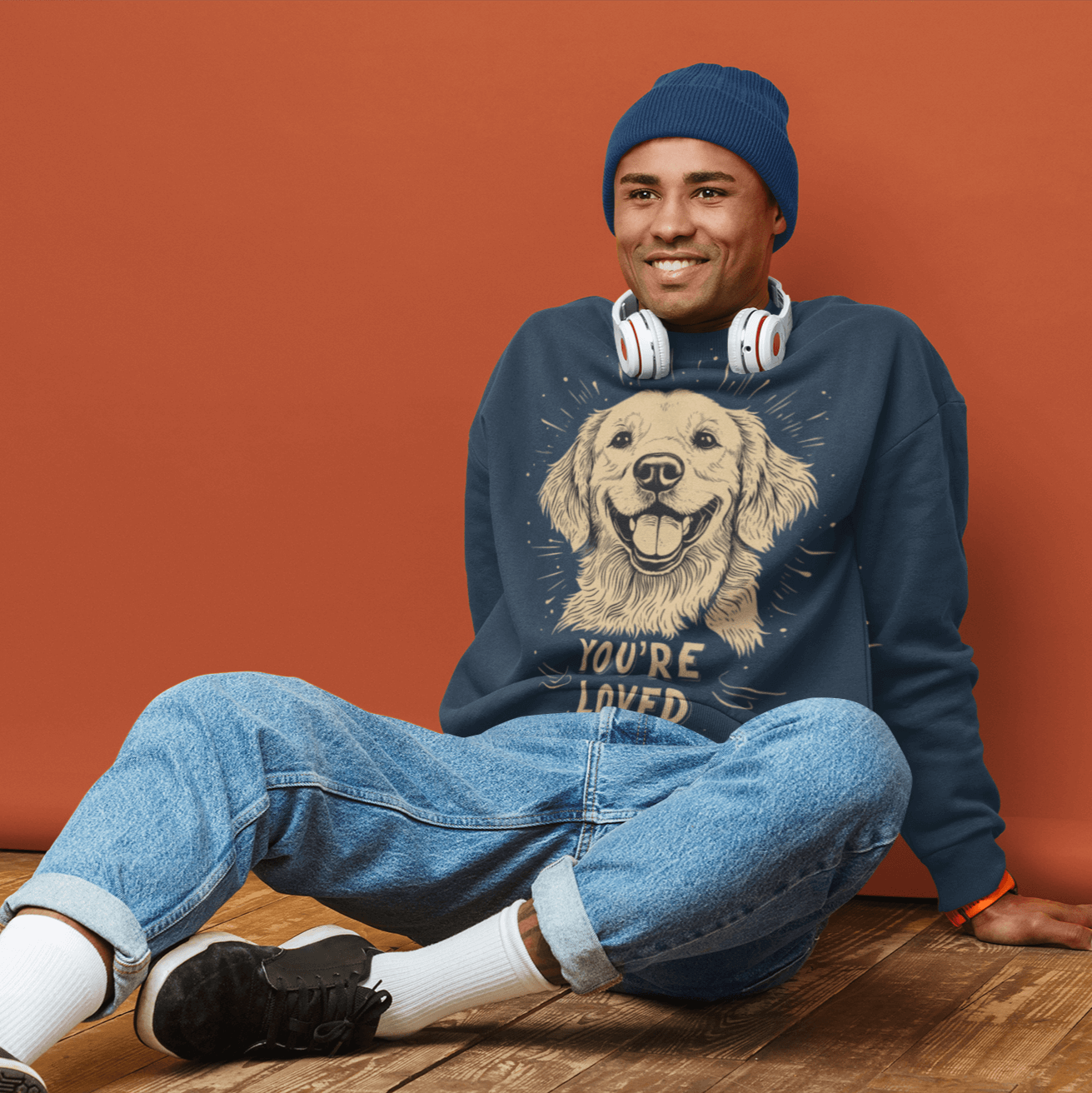 A man wearing a navy blue crewneck sweatshirt sits on a wooden floor with an orange background. The sweatshirt features an illustration of a smiling golden retriever and the text 