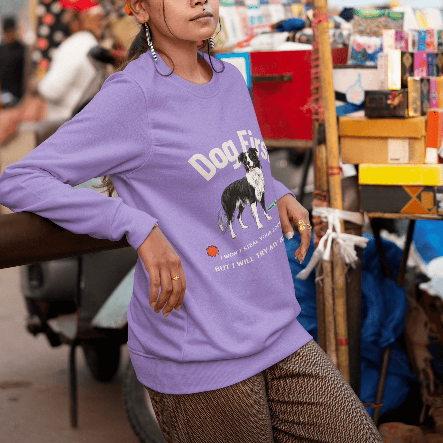 Female model standing by a street market stall, wearing a purple crewneck sweatshirt featuring the text "Dog First" and an illustration of a Border Collie, paired with an orange baseball cap, surrounded by a vibrant market setting.