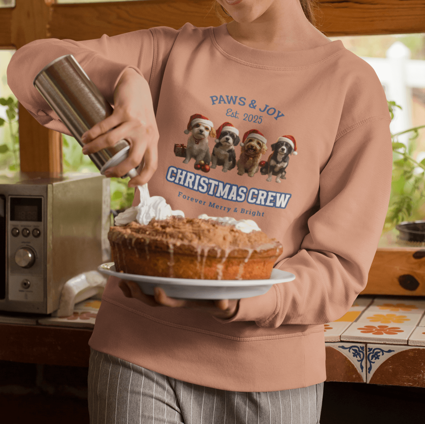 Woman in a Santa hat wearing a pink sweatshirt with "Christmas Crew" and dog illustrations, decorating a Christmas pie with whipped cream in a cozy kitchen setting.