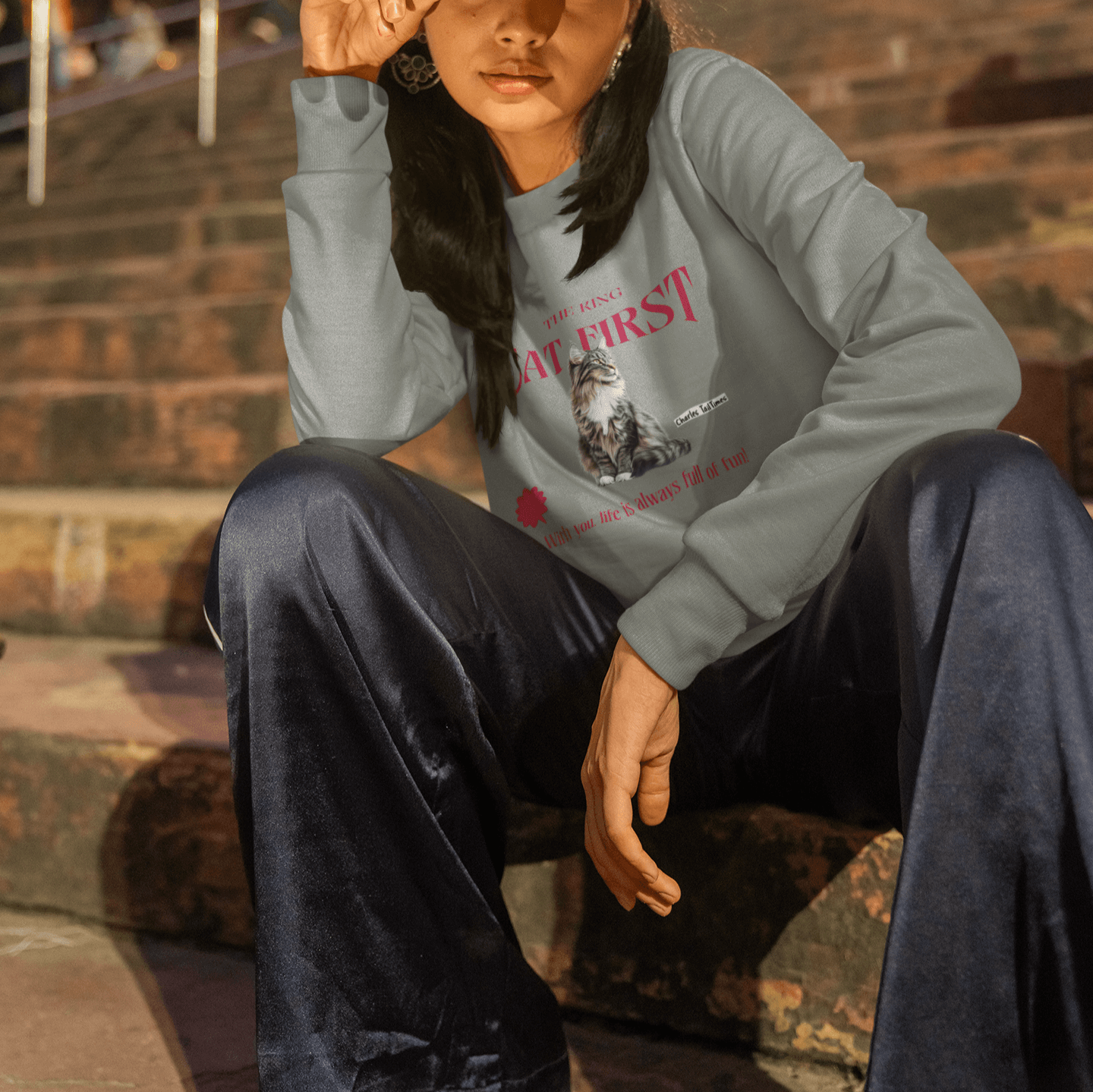 A woman sitting on steps at night, wearing a gray sweatshirt featuring a Maine Coon cat illustration. The sweatshirt has the text "The King" and "Cat First" in bold red, along with the phrase "With you, life is always full of fun!" and Charles TailTimes b