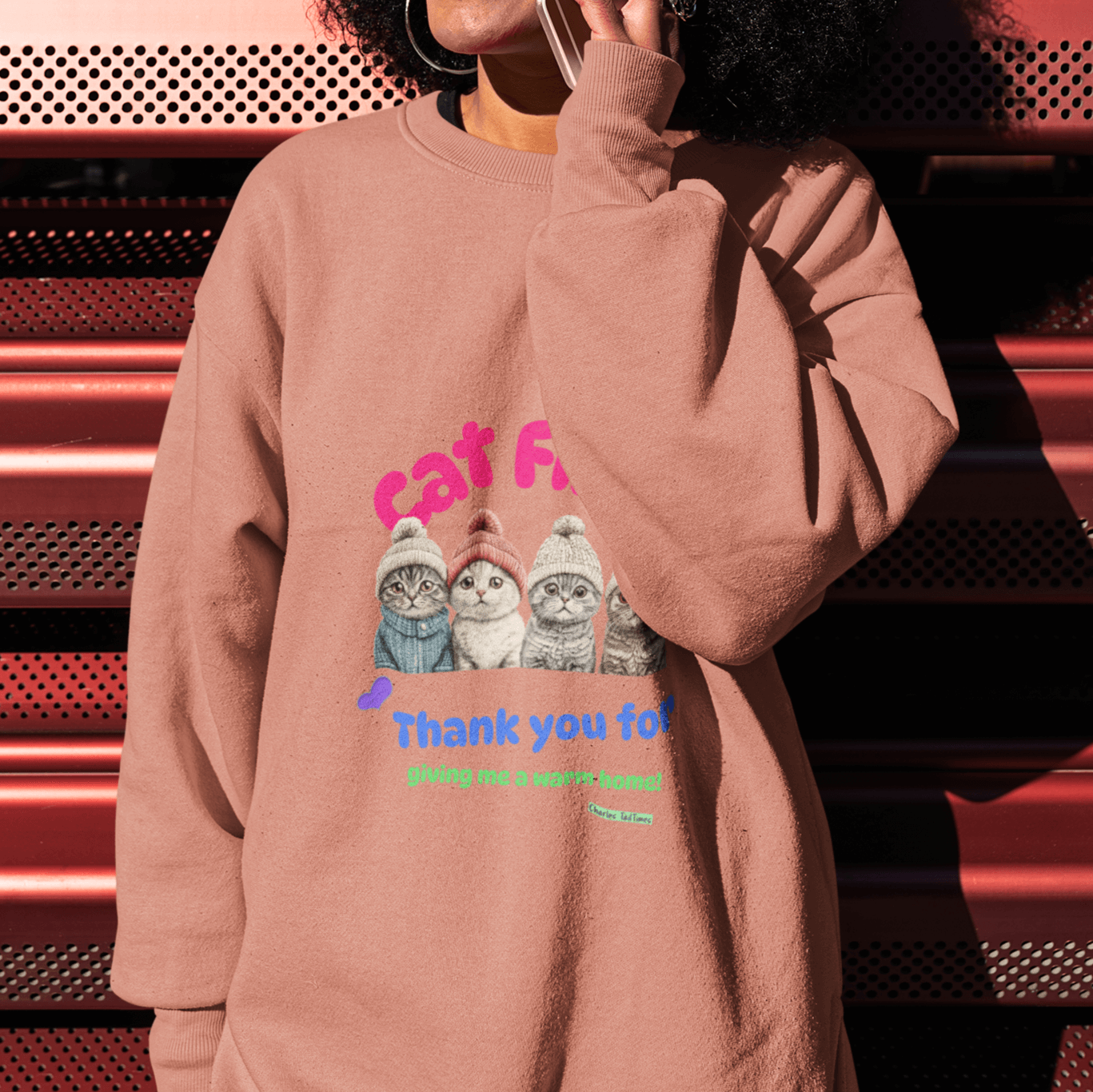 Smiling woman with afro hairstyle wearing a light brown sweatshirt featuring an illustration of four Scottish Fold cats in knitted hats and colorful text 'Cat First' and 'Thank you for giving me a warm home!' while talking on the phone against a red backg