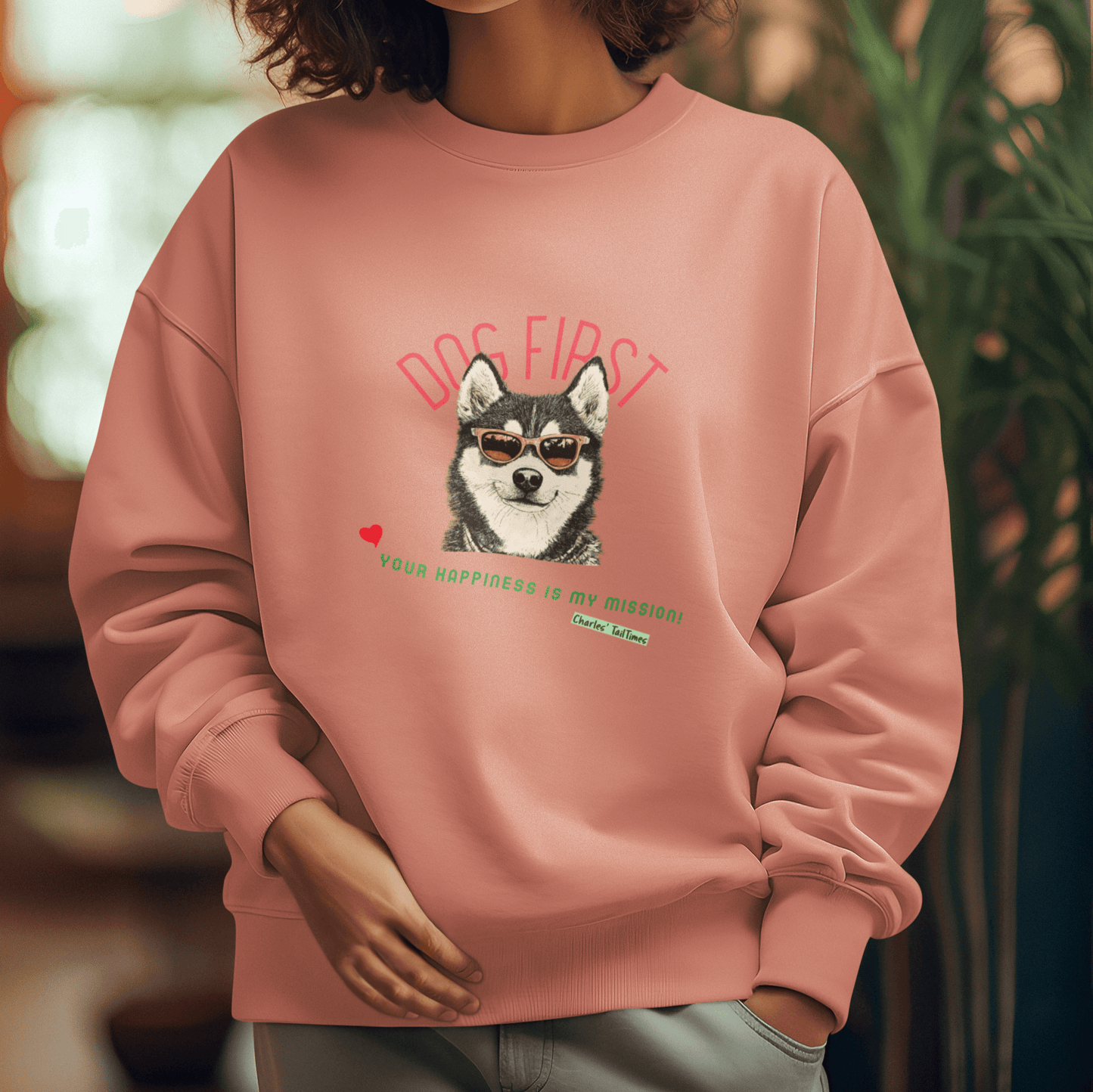 Young female model wearing a pink crewneck sweatshirt featuring an illustration of a Husky wearing sunglasses, with the text "Dog First" in red above and "Your happiness is my mission!" in green below, set against a cozy indoor setting with greenery.