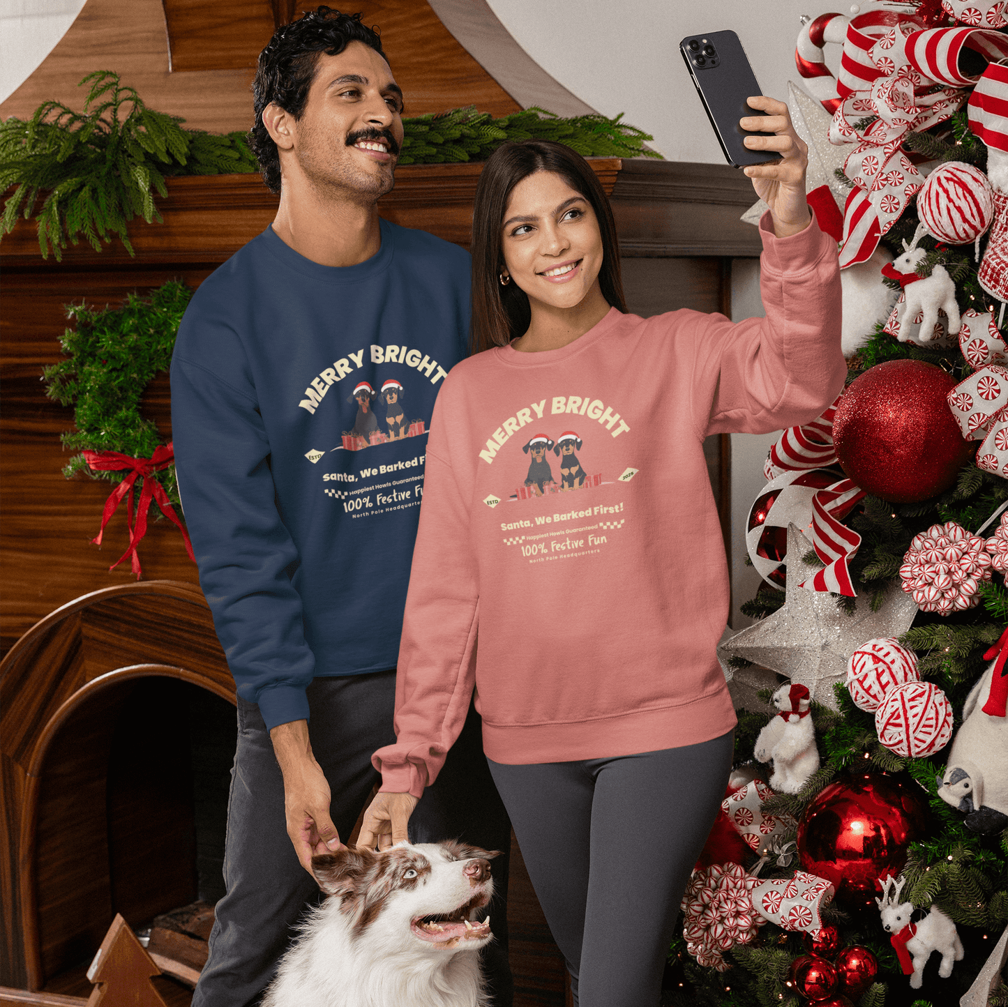 A couple wearing Christmas-themed sweatshirts pose with their shepherd dog in a Christmas-decorated room. The woman is wearing a pink sweatshirt, and the man is wearing a blue sweatshirt, with a Christmas tree and fireplace in the background, creating a w