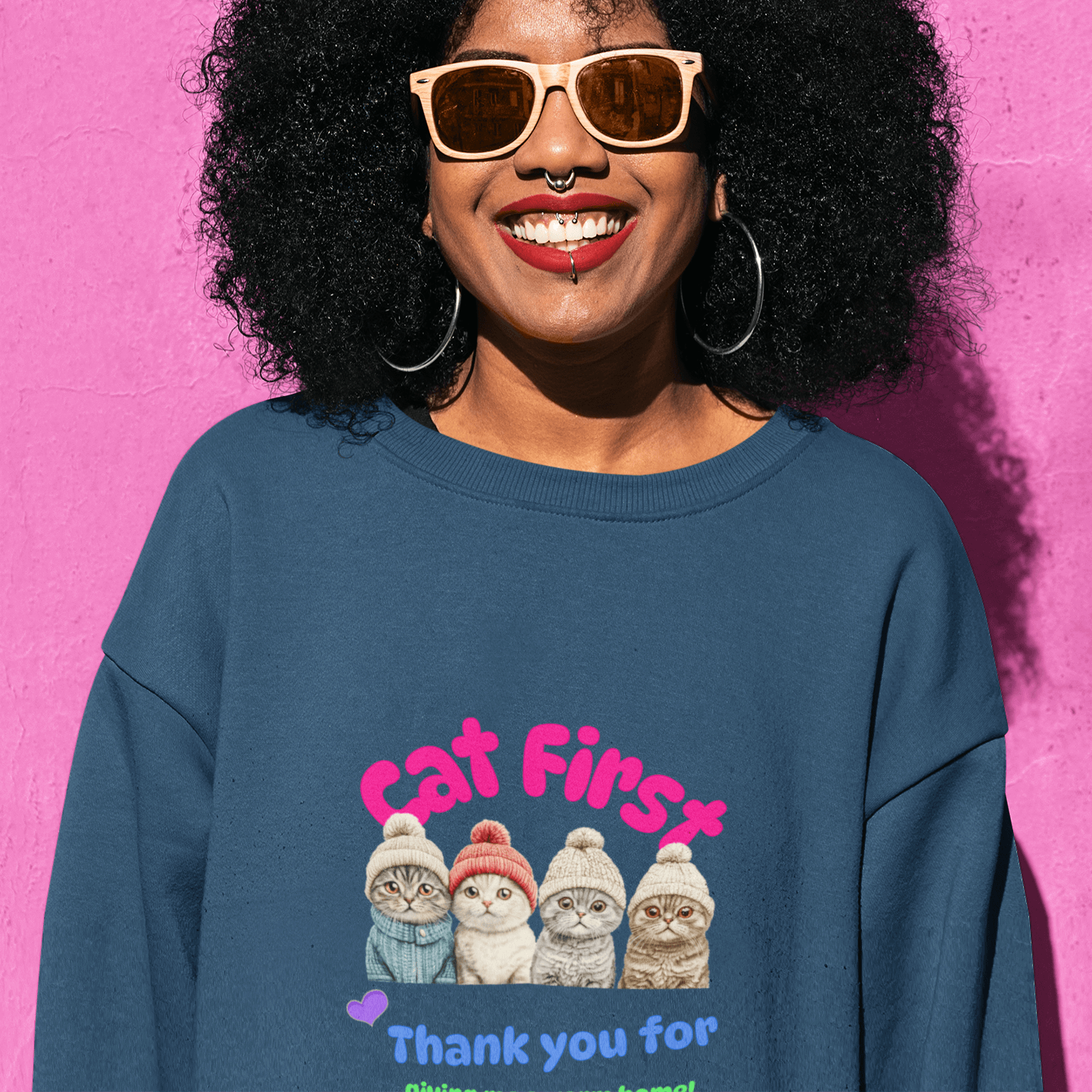 Smiling woman with large curly hair and wooden sunglasses wearing a navy blue sweatshirt featuring a colorful design of four Scottish Fold cats in knitted hats, alongside the text 'Cat First' and 'Thank you for giving me a warm home!' standing against a b