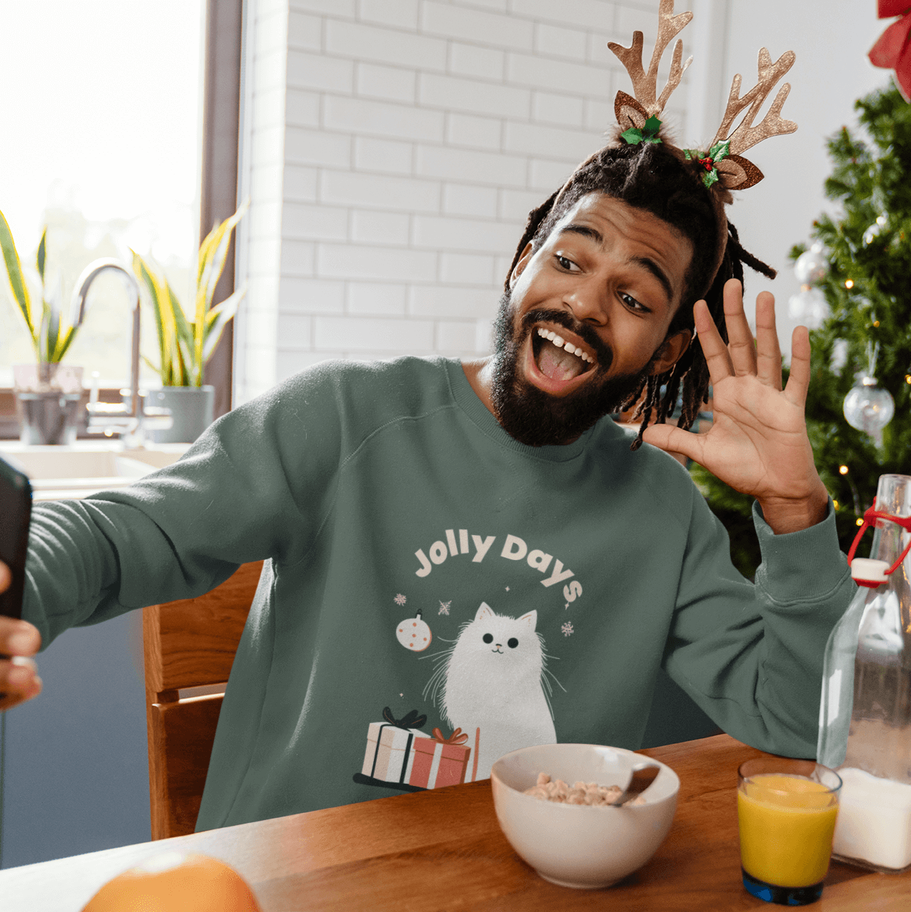 A young man wearing a green Christmas-themed sweatshirt and a reindeer antler headband takes a cheerful selfie at the breakfast table next to a Christmas tree. The sweatshirt features the phrase "Jolly Days" and an illustration of a white cat. The table i