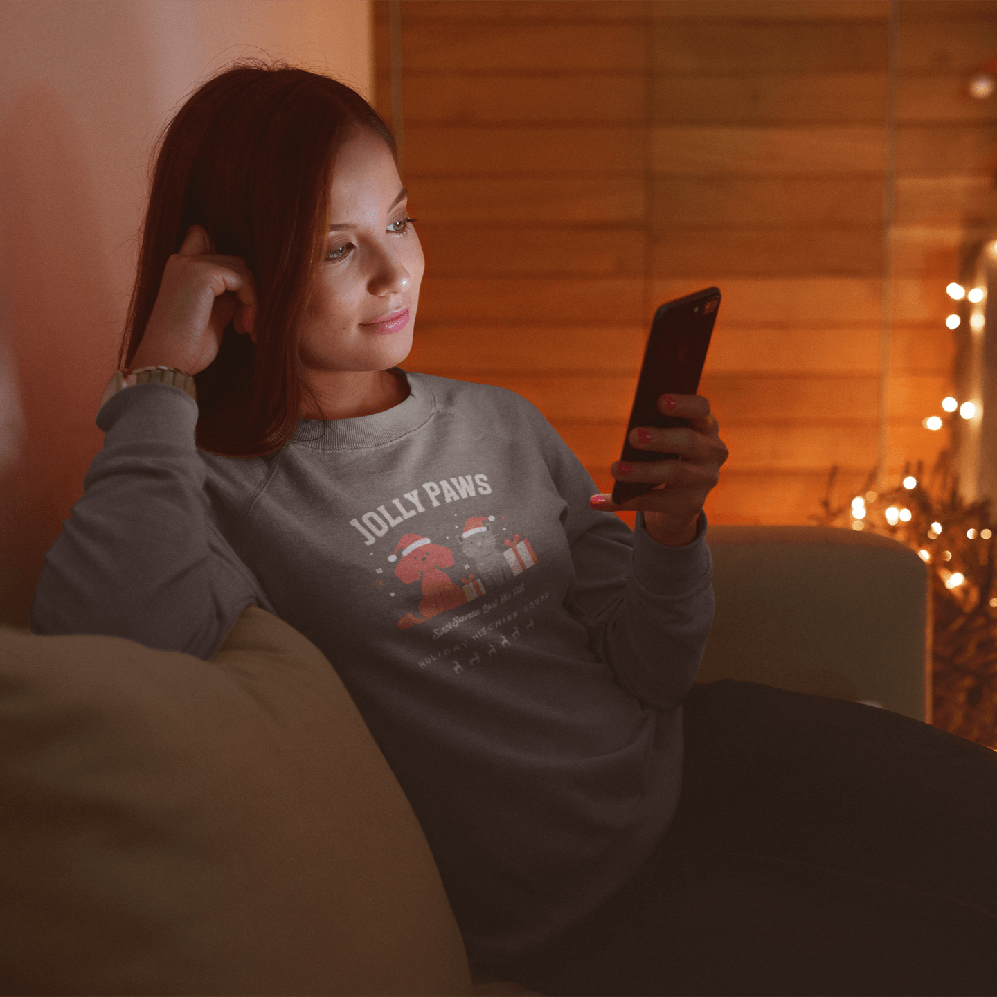 Woman in a cozy sweatshirt with "Jolly Paws" design, relaxing on a couch while using a smartphone during the holidays.