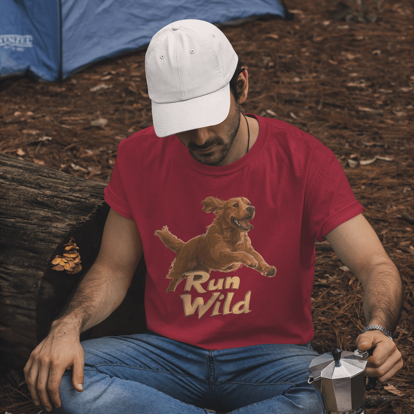 A man sitting at a campsite wearing a red T-shirt with a "Run Wild" design featuring a running Golden Retriever. He is holding a coffee pot, surrounded by camping coffee equipment, with a blue tent in the background, evoking themes of outdoor adventure an