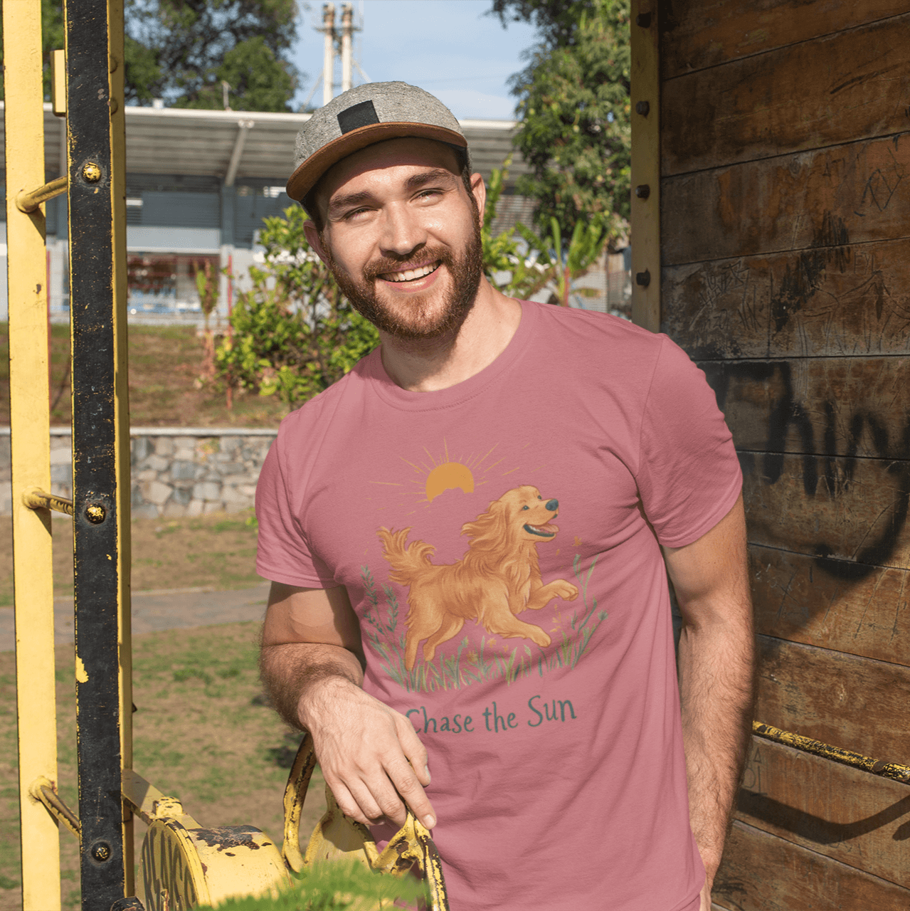 A bearded man wearing a "Chase the Sun" illustration T-shirt, smiling while standing next to a wooden structure in the park, exuding a relaxed and casual vibe.