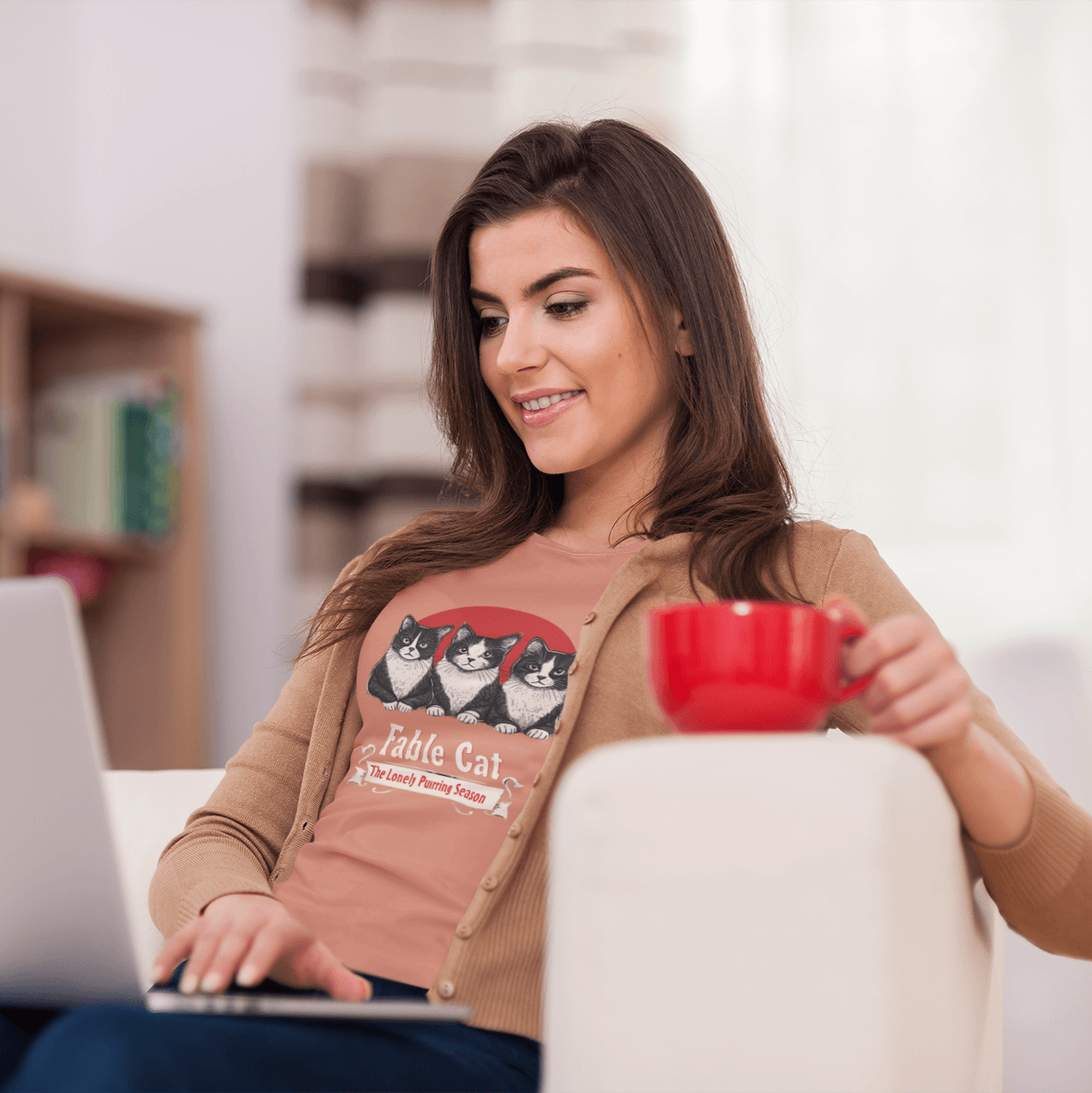 Smiling woman working on a laptop, wearing a peach T-shirt with 'Fable Cat' design featuring three cats and a red backdrop, holding a red coffee mug.