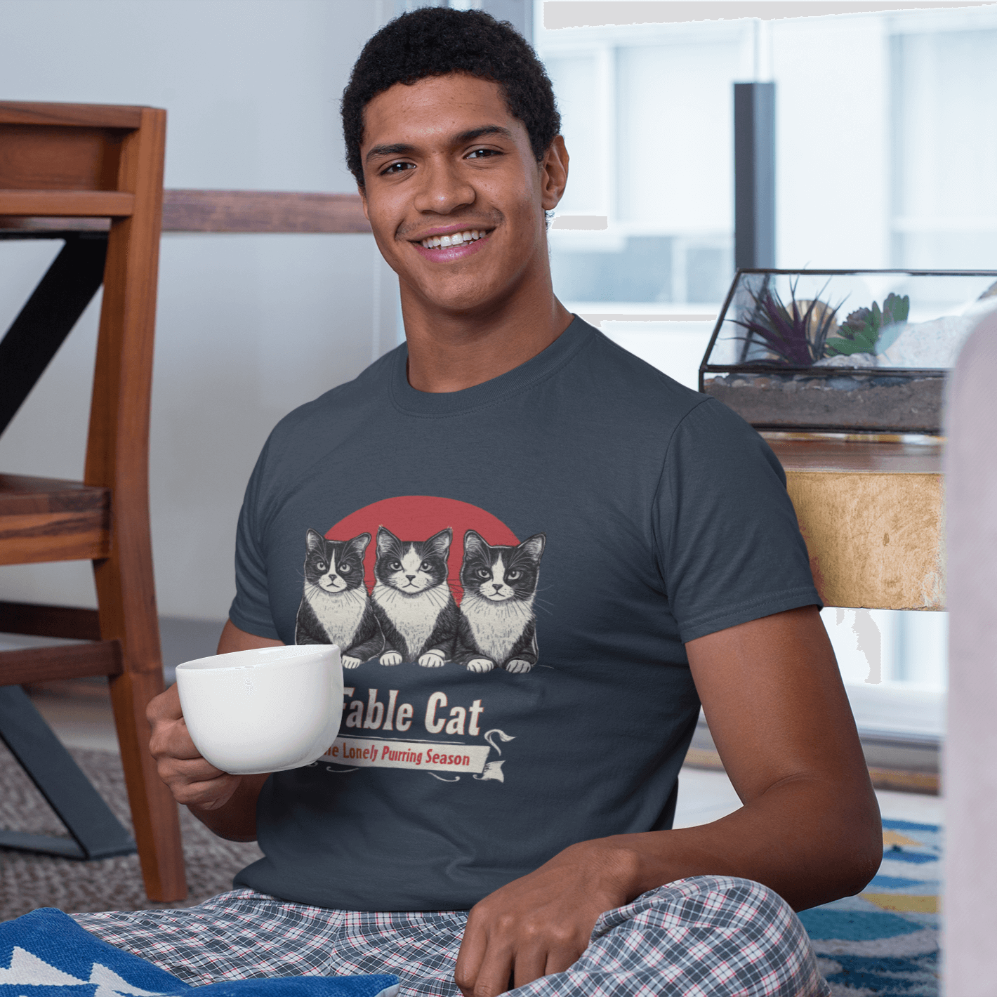 Smiling man sitting in a living room holding a white mug, wearing a dark T-shirt featuring a design of three cats with the text 'Fable Cat: The Lonely Purring Season.