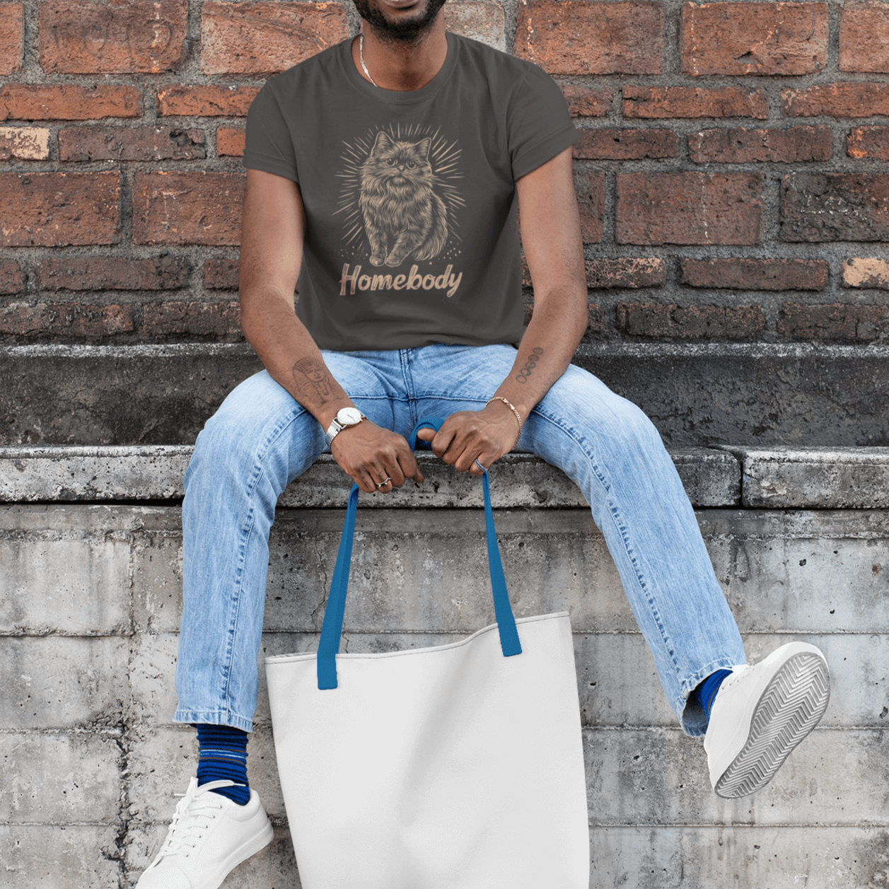 A model sitting in front of a brick wall wearing a gray T-shirt with a Persian cat illustration and the text 'Homebody,' holding a white tote bag, paired with blue jeans and white sneakers, showcasing a casual style.