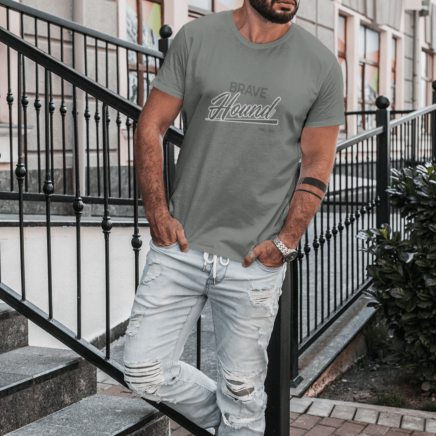 A man wearing a muted green T-shirt with a 'Brave Hound' graphic design, standing on outdoor concrete steps near a black metal railing, styled with distressed light-wash jeans and a confident expression.